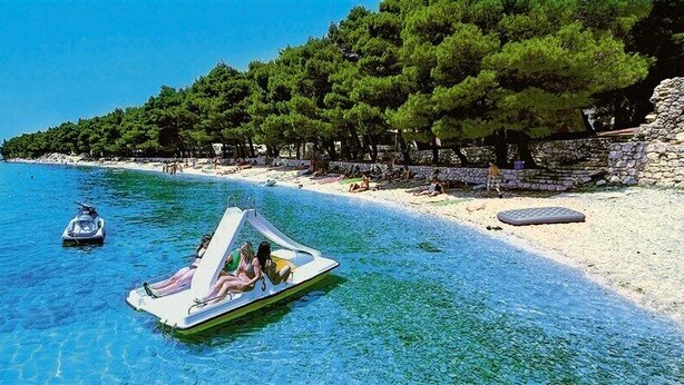 A clear blue sea with a boat in the foreground and a beach with trees and a stone wall in the background. - Victoria Mobilehome Camping Dole (Podgora)