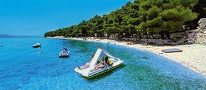 A clear blue sea with a boat in the foreground and a beach with trees and a stone wall in the background. - Victoria Mobilehome Camping Dole (Podgora)