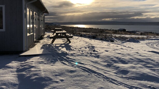 A wooden building with a covered patio area, a picnic table, and a snowy landscape with a body of water in the background. - Hvammstangi Hill Homes (Hvammstangi)