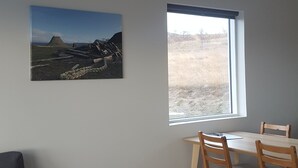 A room with a dining table and chairs, a gray sofa, a painting of a landscape, and a window with a view of a field. - Hvammstangi Hill Homes (Hvammstangi)