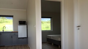 A compact kitchen with a refrigerator, sink, and window. A bed is visible in the adjacent room. - Hvammstangi Hill Homes (Hvammstangi)