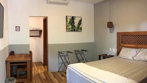 Ein Schlafzimmer mit einem hölzernen Bett, einem kleinen Schreibtisch und einem Stuhl. Ein Wäscheständer und ein Regal mit Gegenständen sind an der Wand angebracht.