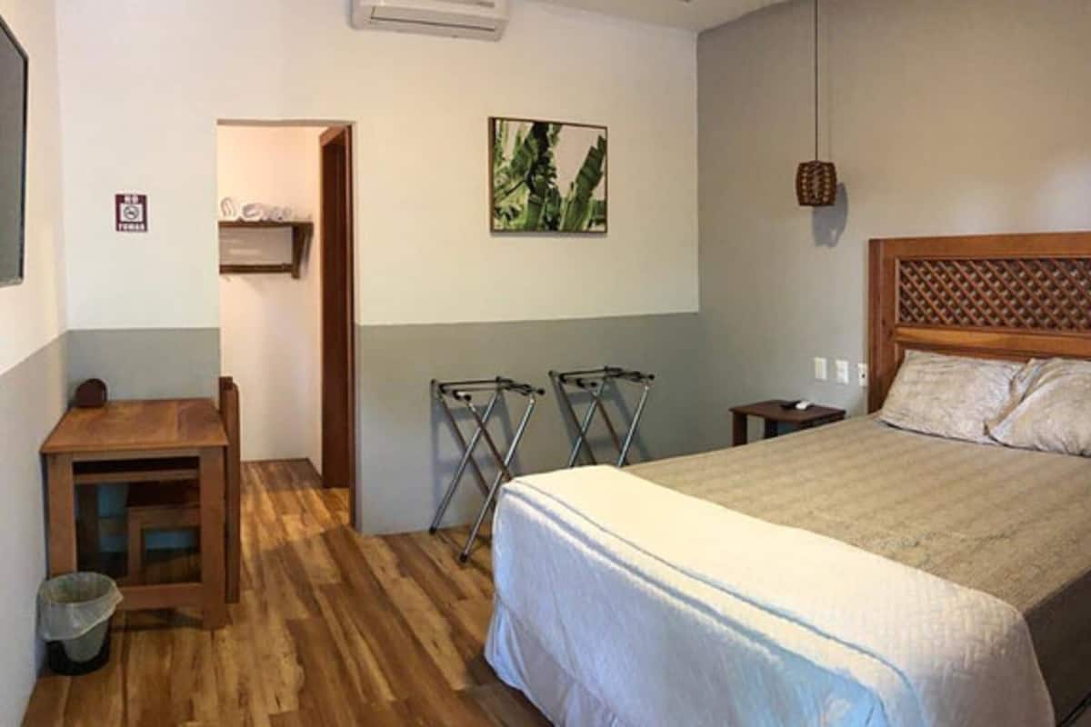 A bedroom with a wooden bed, a small desk, and a chair. There is a drying rack and a shelf with items on the wall.