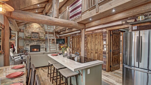 A rustic cabin interior with a stone fireplace, wooden beams, a large kitchen island, and a stainless steel refrigerator. - SkyRun Cabin - Stunning Mountain Views & Fire Pit (Golden)