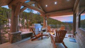 A covered outdoor area with a grill, wooden chairs, and a view of a forested landscape. - SkyRun Cabin - Stunning Mountain Views & Fire Pit (Golden)