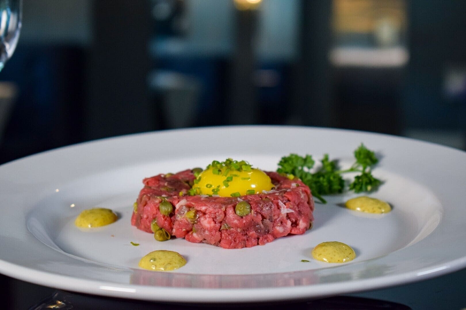 Un plato de tartare de ternera con yema de huevo cruda, adornado con cebollín y perejil, servido en un plato blanco.