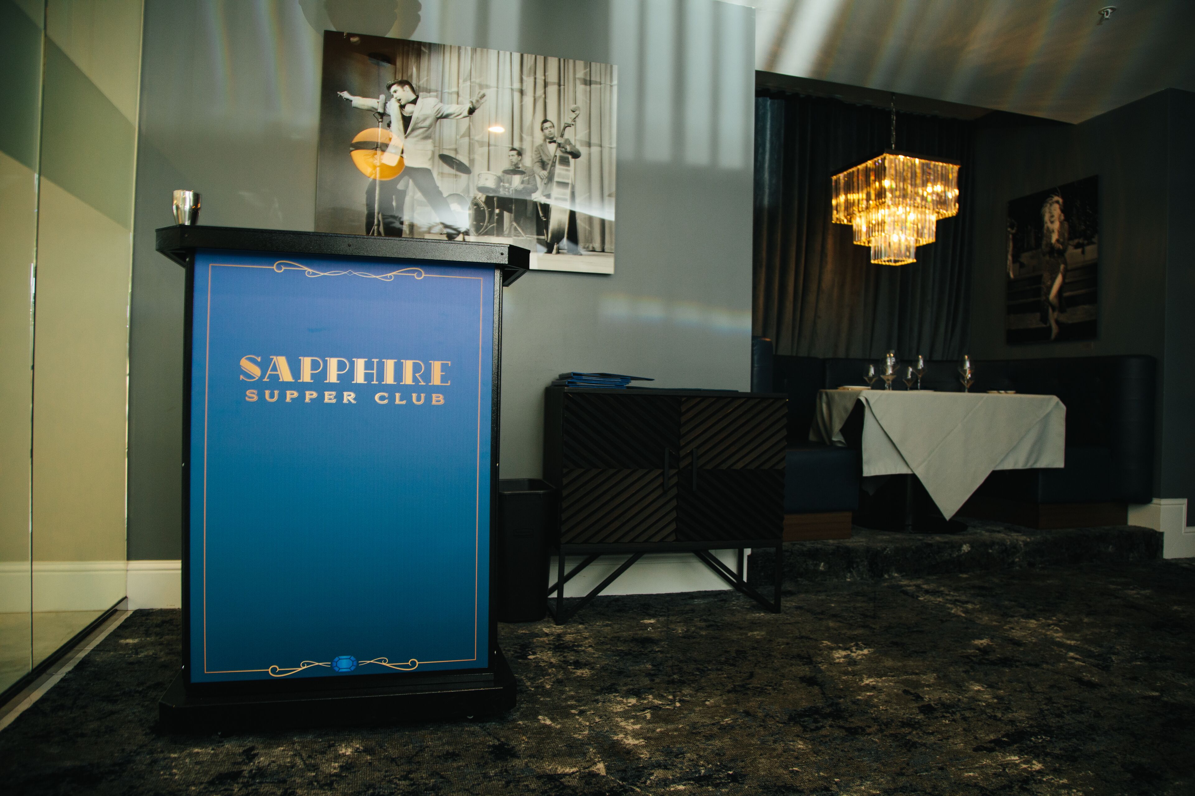 A room with a blue sign reading "Sapphire Supper Club," a chandelier, a table with a white tablecloth, and a framed picture on the wall.