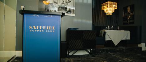 A room with a blue sign reading "Sapphire Supper Club," a chandelier, a table with a white tablecloth, and a framed picture on the wall.