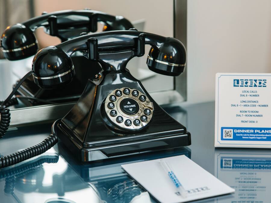 Un teléfono rotativo negro antiguo sobre una mesa de vidrio, junto con un bloc de notas y un bolígrafo, y un cartel que detalla los servicios del hotel.