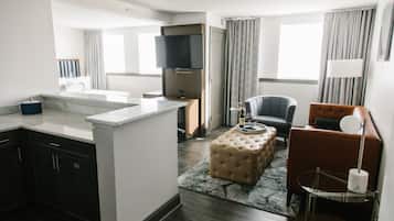 Une chambre d’hôtel moderne dotée d’une kitchenette, d’un téléviseur à écran plat, d’un coin salon confortable avec un canapé et un fauteuil, et d’une grande fenêtre avec des rideaux.
