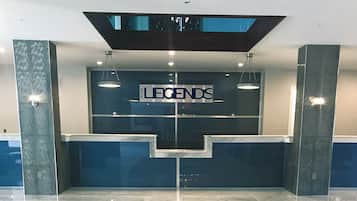 Uma área de recepção de hotel com design moderno, com um balcão de atendimento, um relógio digital e uma placa com a inscrição 'LEGENDS'.