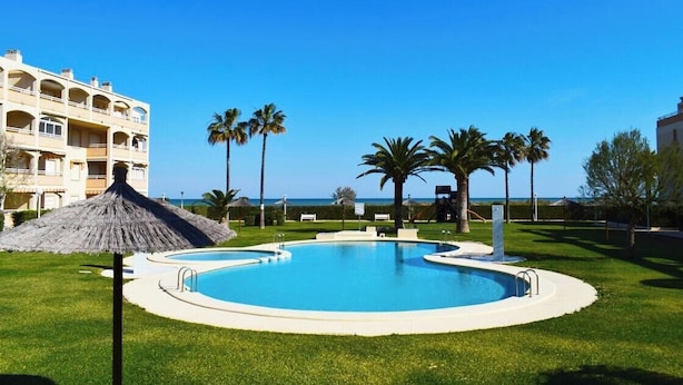 A swimming pool with a thatched umbrella, surrounded by palm trees and a building with balconies. - Anacasa Bahia de Denia Les Marines AP2102 (Denia)