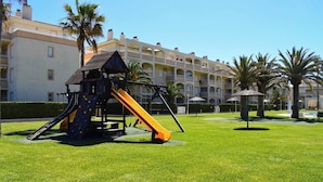 A playground with a slide and climbing structure in front of a hotel with multiple balconies and palm trees. - Anacasa Bahia de Denia Les Marines AP2102 (Denia)