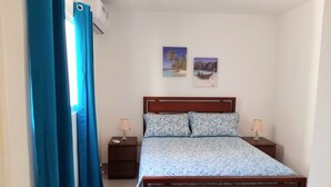 A bedroom with a wooden bed, two bedside tables with lamps, a window with blue curtains, and two wall-mounted pictures. - Summer paradise, total relaxation on your vacation (Stgo)