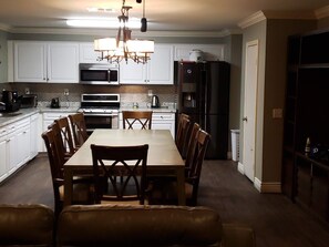 A modern kitchen with white cabinets, a dining table with chairs, a chandelier, and a refrigerator.