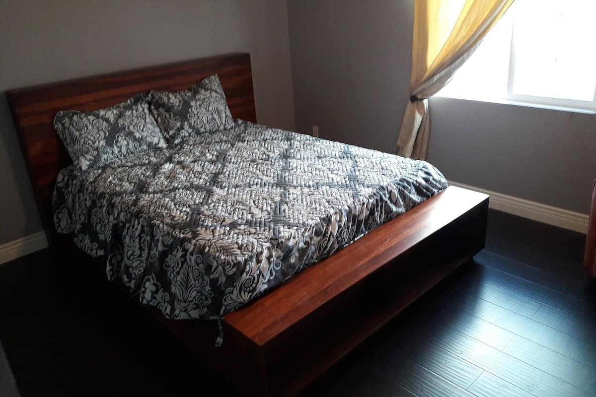 A bed with a patterned bedspread in a room with a wooden floor and a window with curtains.
