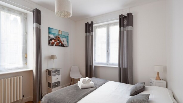 A modern bedroom with a bed, bedside tables, a lamp, a chair, and a window with curtains. - Le Hauban - Three Bedroom Apartment, Sleeps 6 (Saint-Malo)