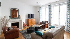 A living room with a fireplace, leather sofas, a television, and patterned curtains. - Le Hauban - Three Bedroom Apartment, Sleeps 6 (Saint-Malo)