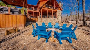 A cabin with a stone exterior, a wooden deck, and a fire pit surrounded by blue Adirondack chairs.