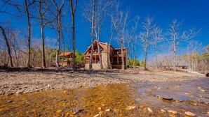 A cabin with a stone foundation and wooden exterior, surrounded by trees and a clear stream.