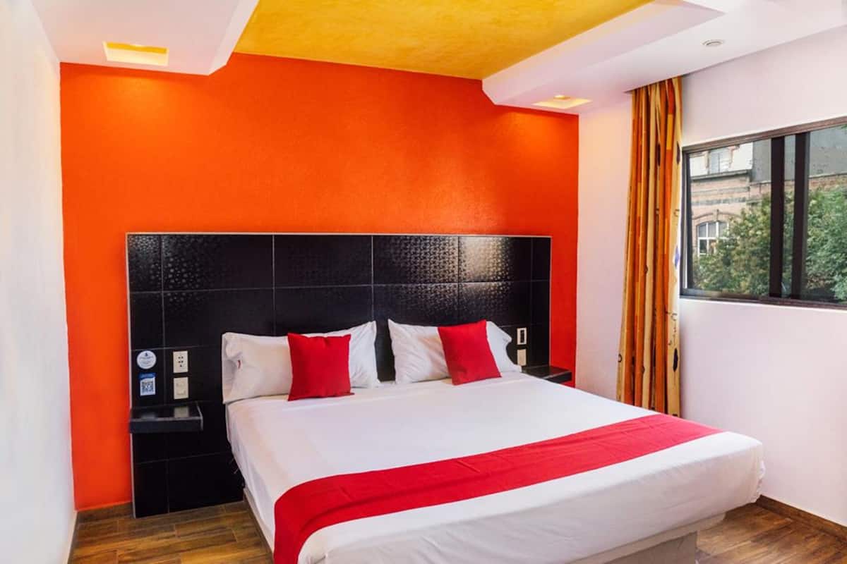 Une chambre d’hôtel avec un grand lit, des murs orange et une vue sur la ville par la fenêtre.