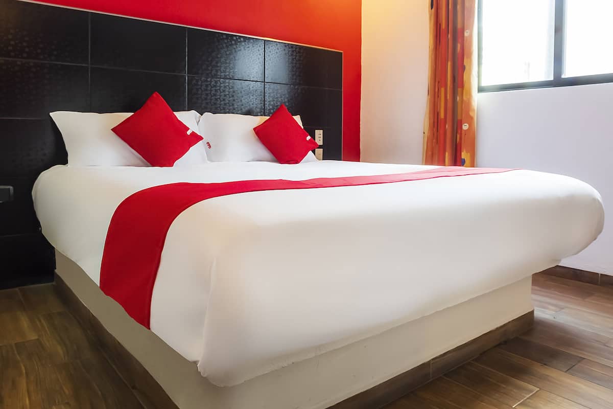 Une chambre d’hôtel moderne avec un grand lit, une literie rouge et blanche, une tête de lit noire et une vue sur l’extérieur par une fenêtre.