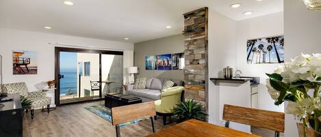 A modern living room with a sofa, coffee table, and dining area. There is a view of the ocean through a sliding door.