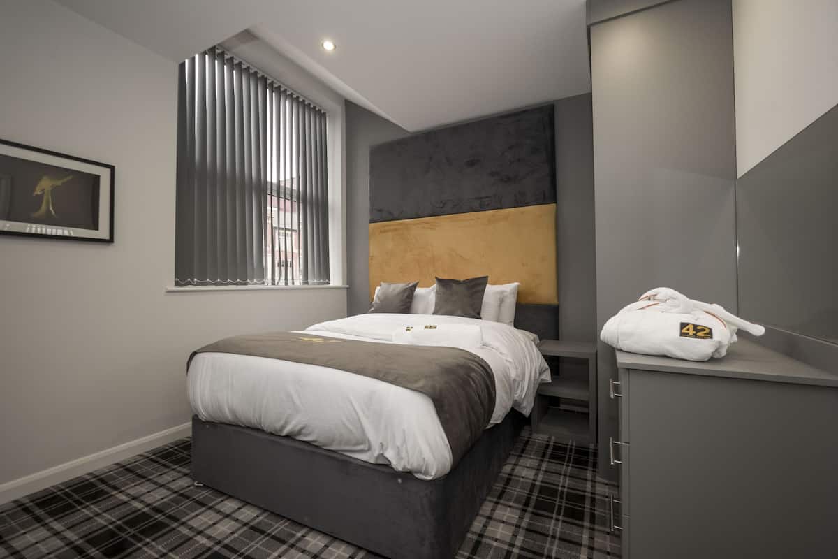 A modern hotel room with a large bed, a headboard with a textured design, a window with blinds, a framed picture on the wall, and a robe hanging on a cabinet.