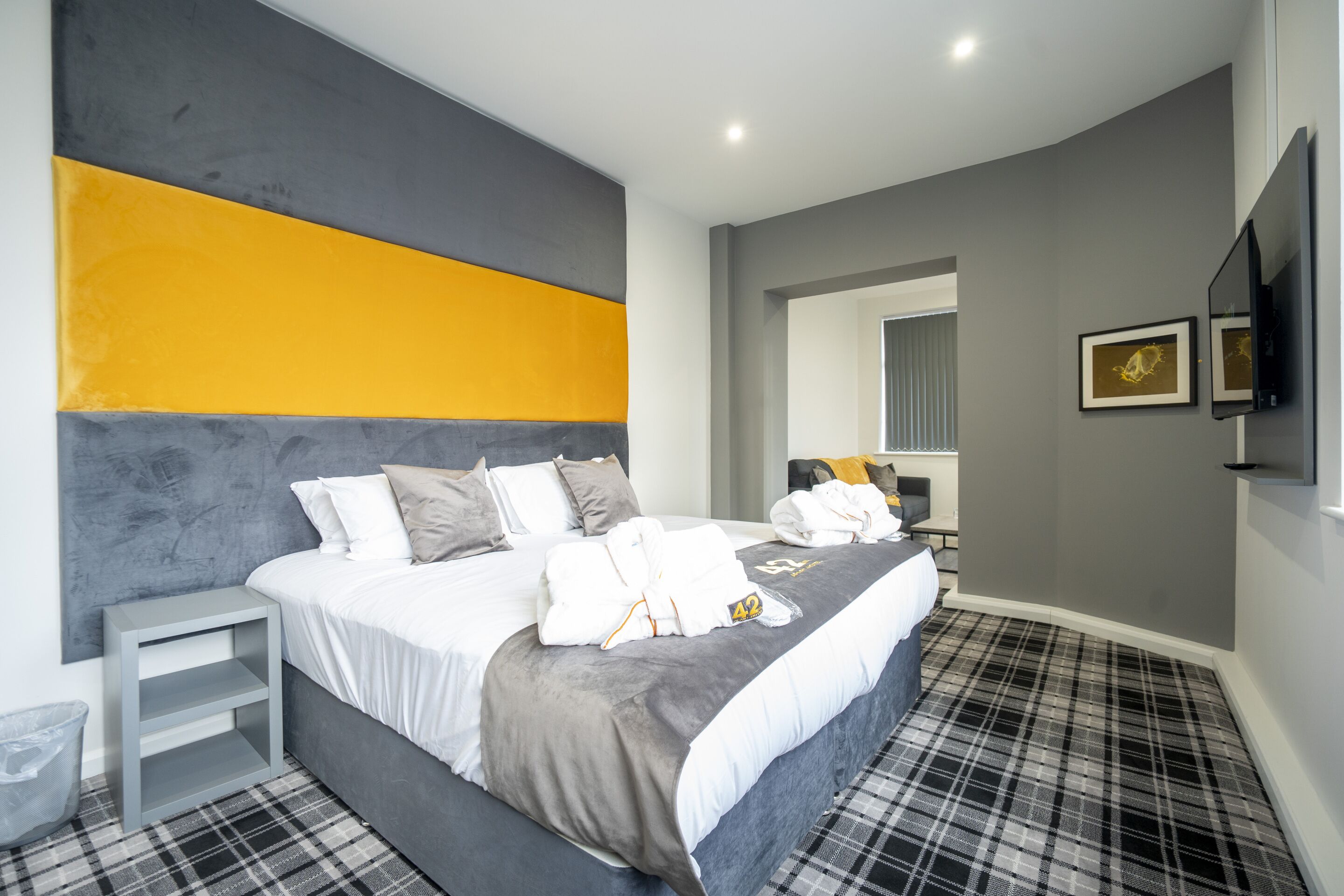 A modern hotel room with a large bed, a grey and yellow accent wall, a flat-screen TV, and a plaid-patterned carpet.