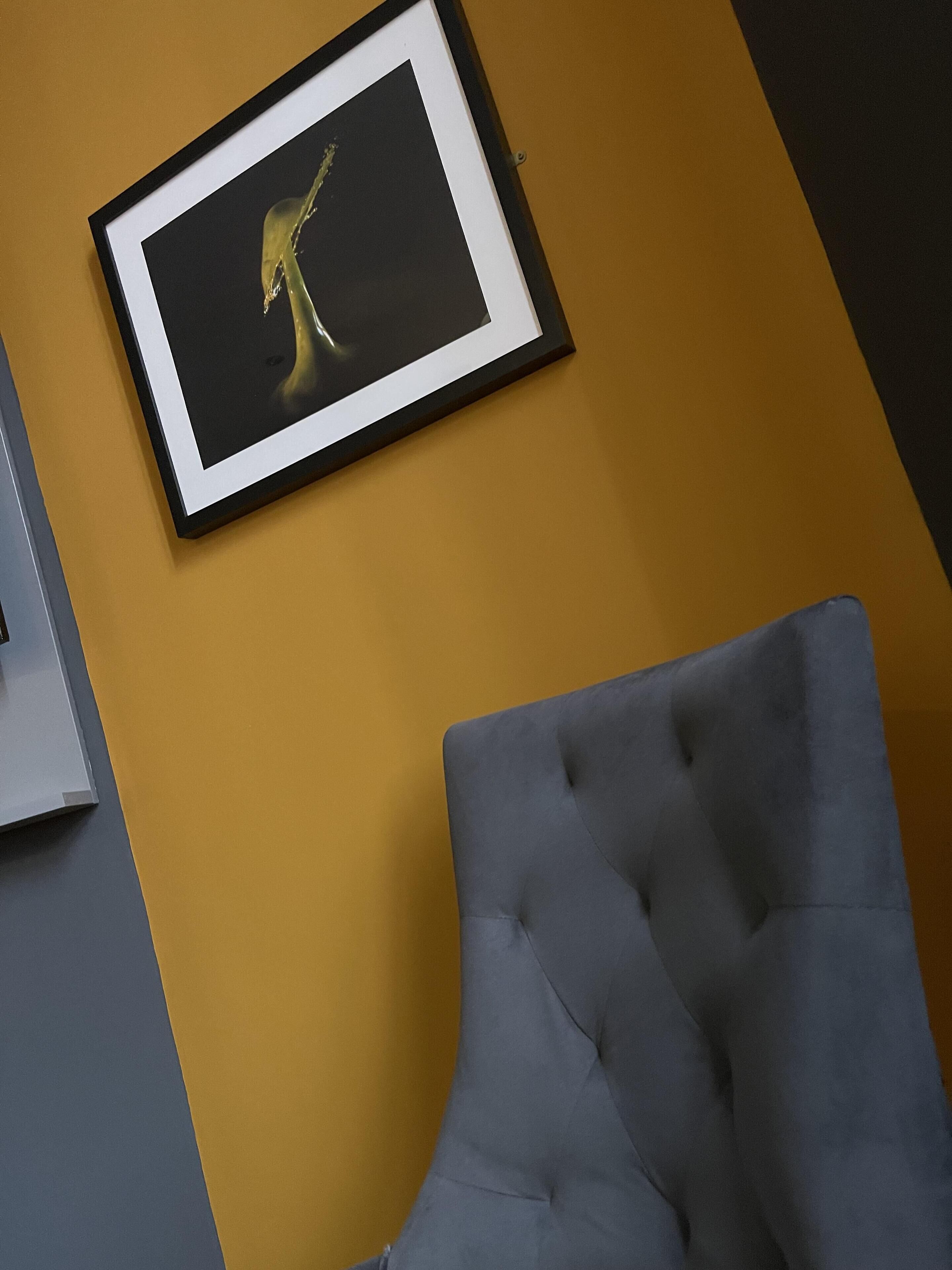 Une œuvre d’art encadrée sur un mur jaune, un fauteuil gris à motifs et un meuble gris.