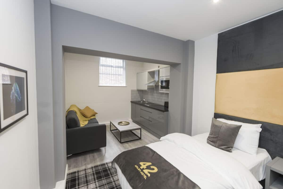 A modern hotel room with a bed, a sofa, a small table, a kitchen area, and a window.