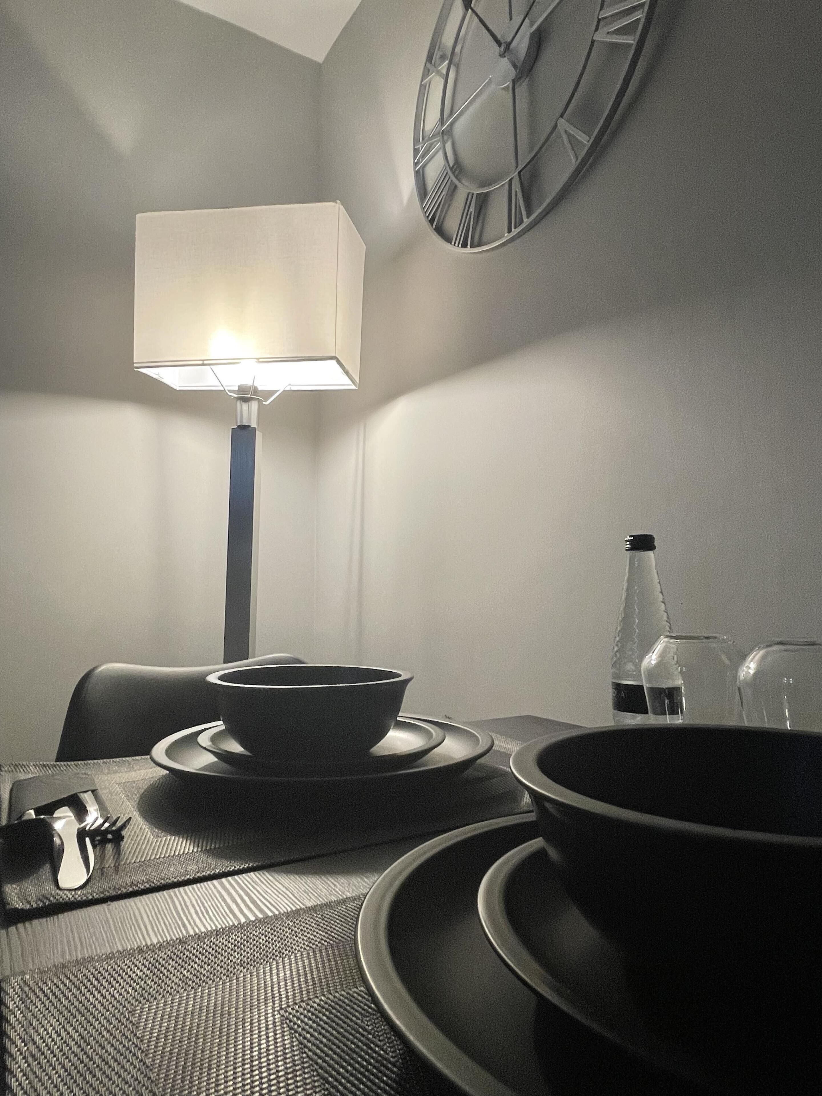 Uma área de refeições com uma mesa posta para dois, um relógio na parede e um abajur.