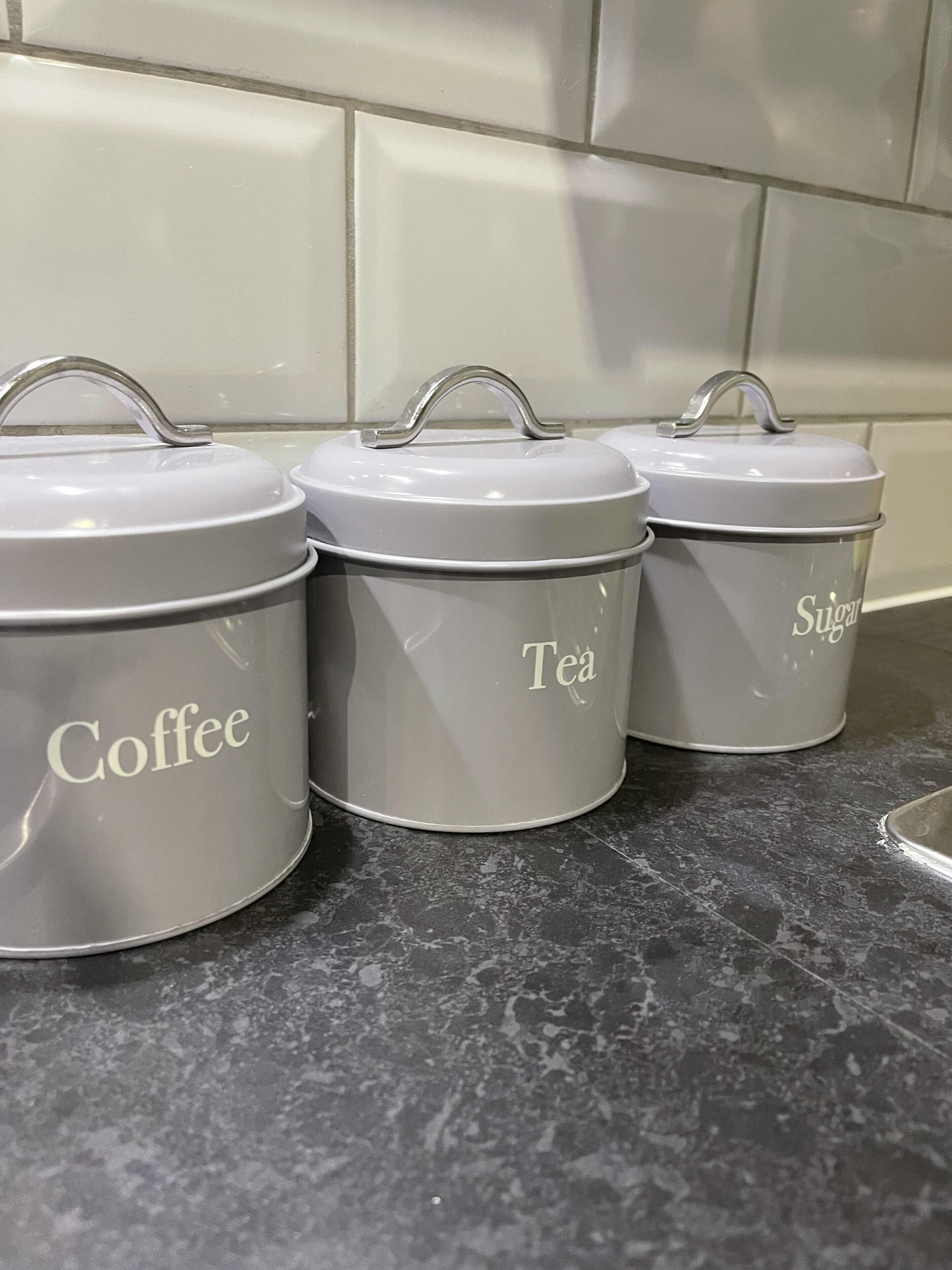 Trois boîtes de rangement grises sont posées sur le plan de travail de la cuisine, et portent les étiquettes suivantes : « Café », « Thé » et « Sucre ».