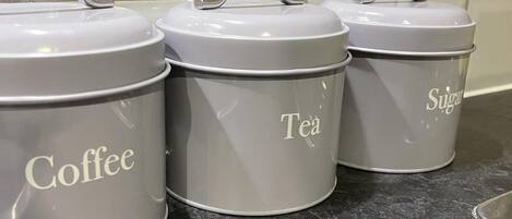 Trois boîtes de rangement grises sont posées sur le plan de travail de la cuisine, et portent les étiquettes suivantes : « Café », « Thé » et « Sucre ».