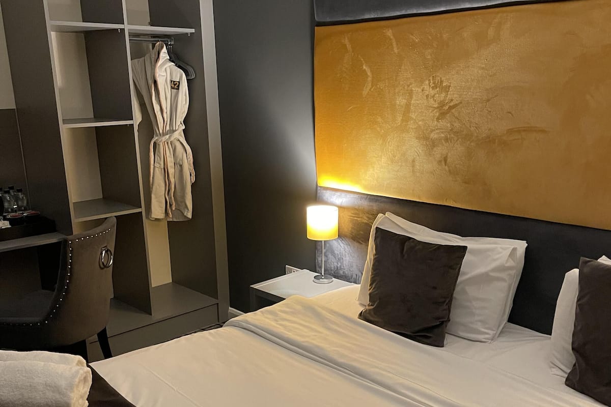A hotel room with a bed, a robe hanging on a rack, a desk with a chair, and a nightstand with a lamp.