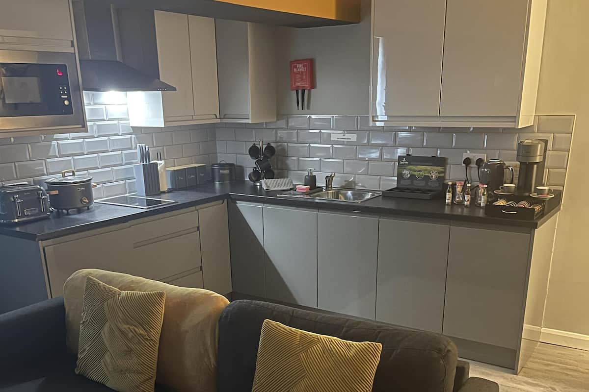 A modern kitchen with grey cabinets, a black countertop, and a built-in microwave. There is a black sofa with two cushions in the living area.