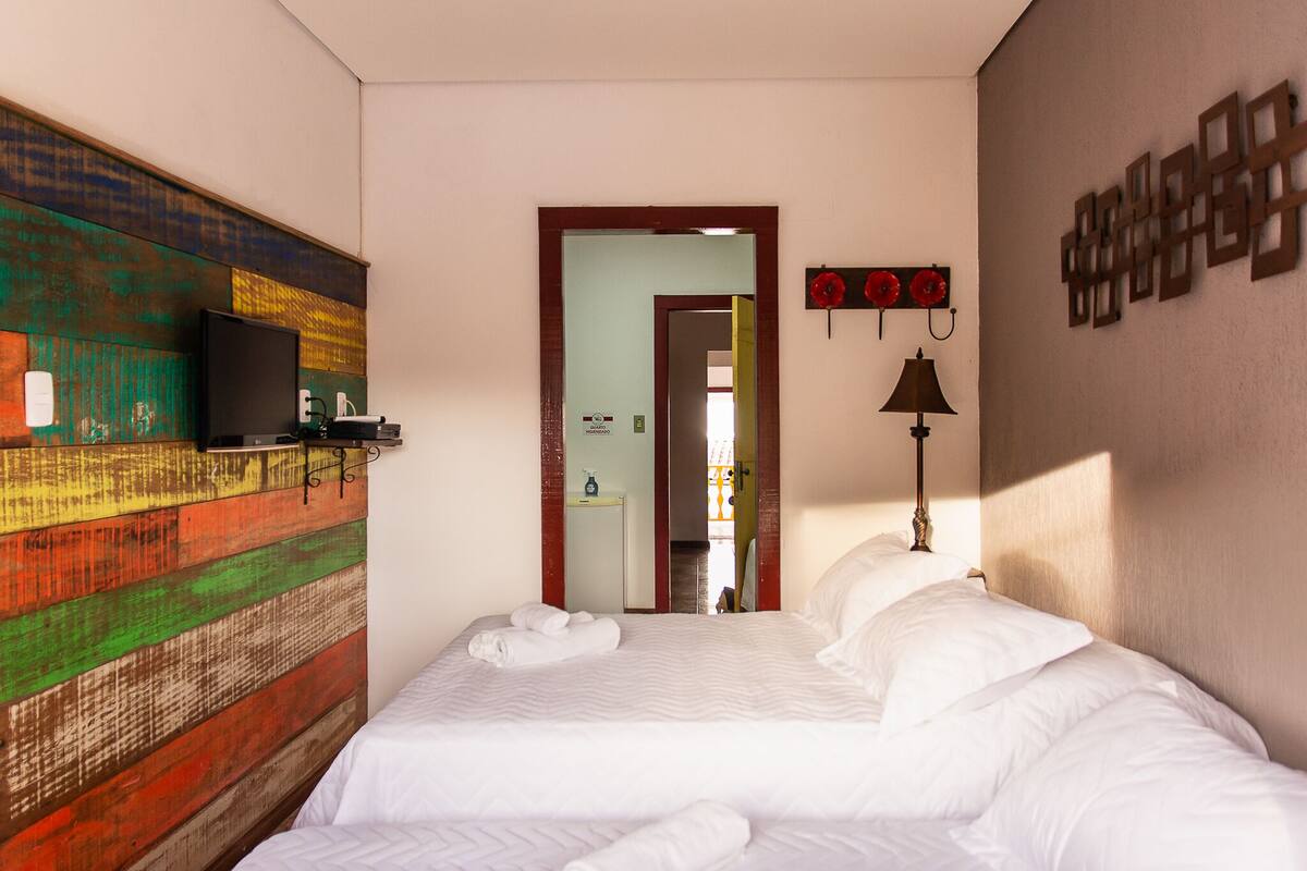 Quarto de hotel com cama, televisão em uma prateleira, espelho e parede com design colorido.