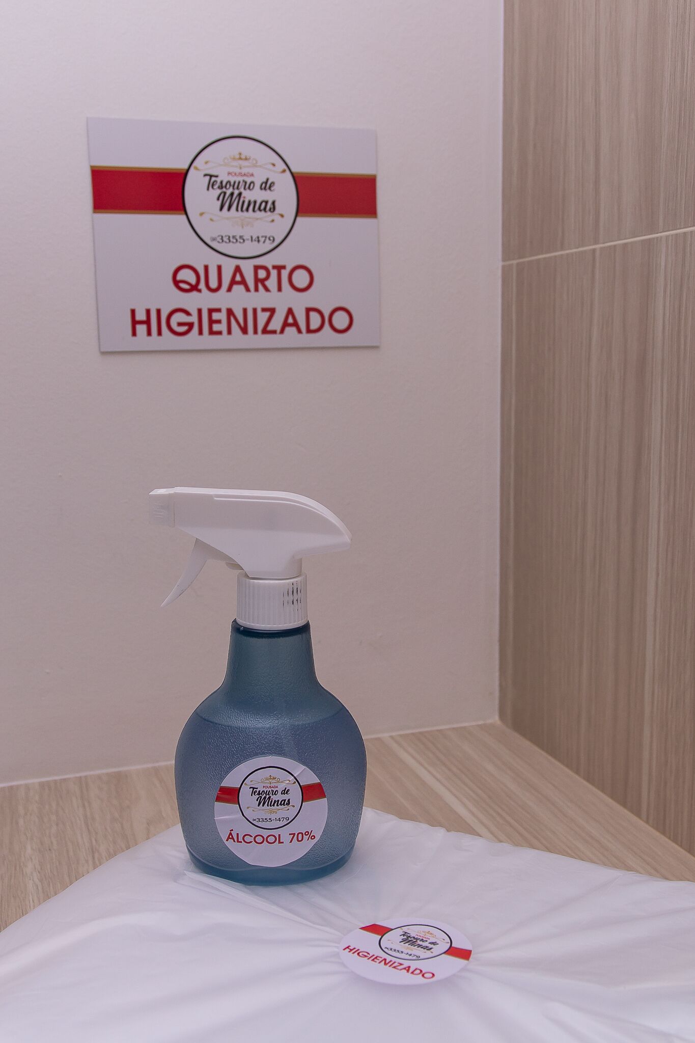 Un flacone spray con l'etichetta 'ÁLCOOL 70%' e un cartello che riporta la scritta 'QUARTO HIGIENIZADO' in una camera d'albergo.