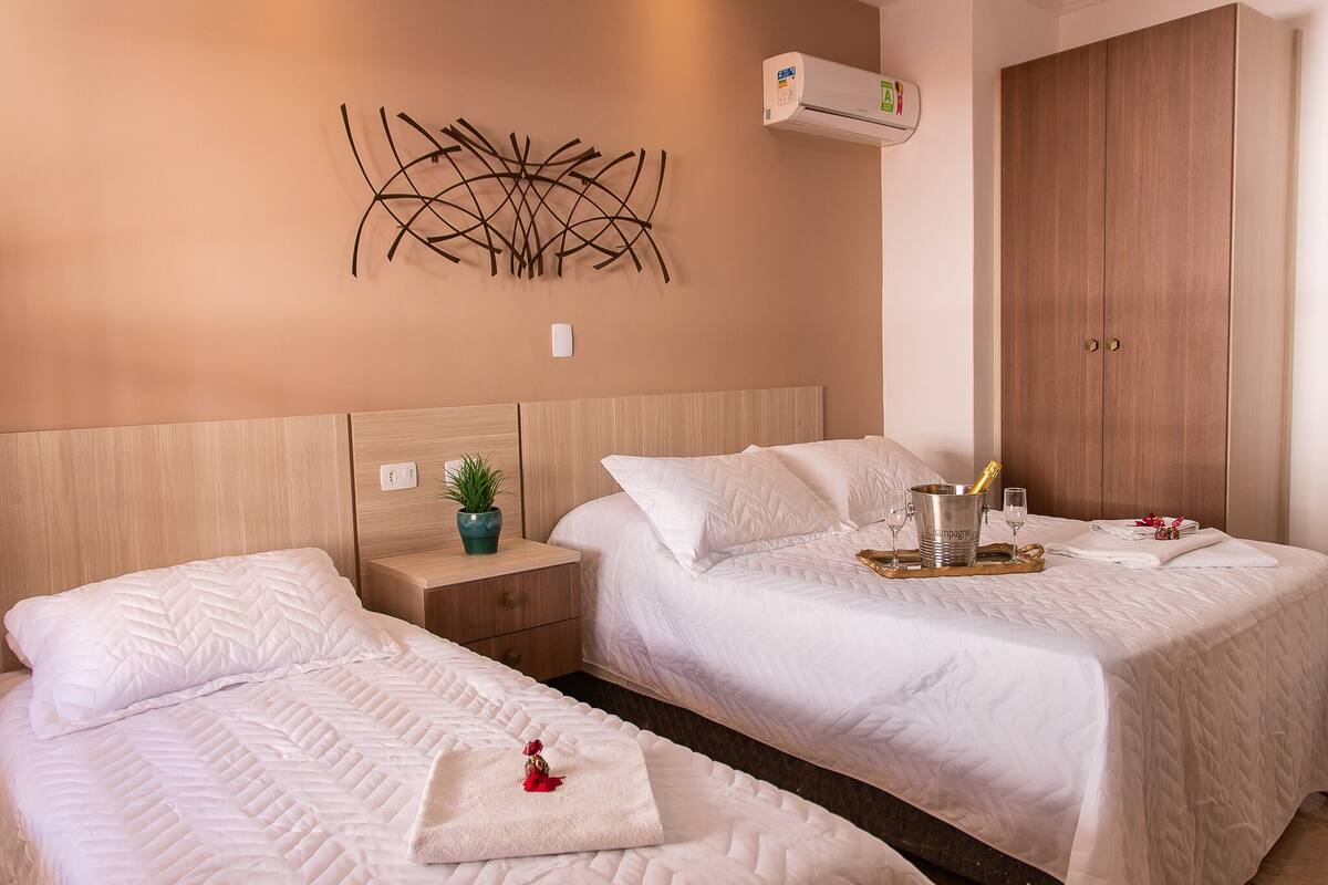 Quarto de hotel com duas camas, cabeceira de madeira, ar condicionado fixado na parede e guarda-roupa de madeira.
