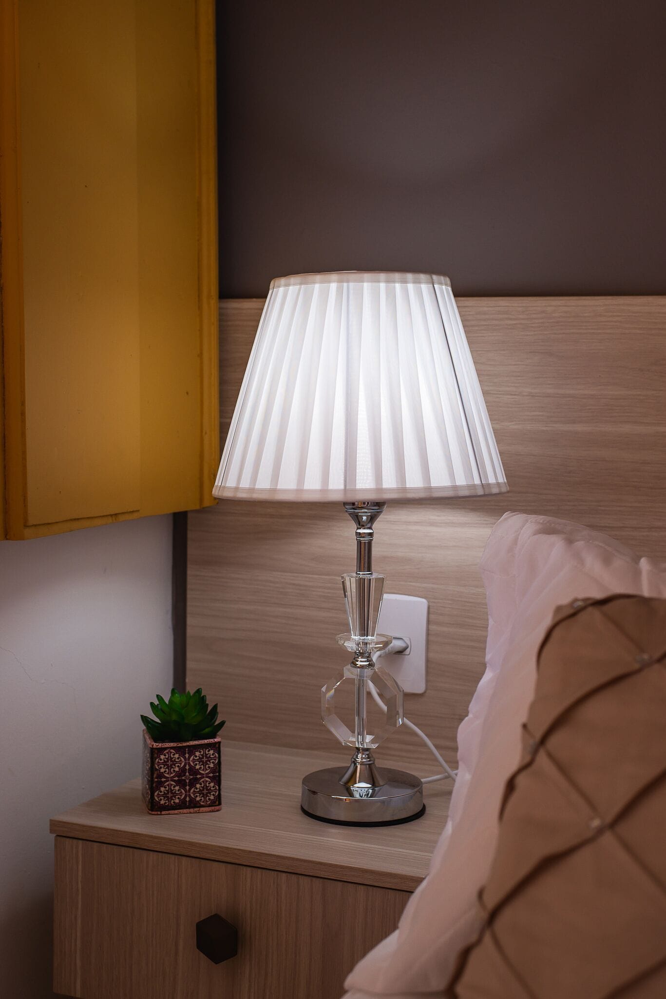 Uma mesa de cabeceira com uma luminária, um vaso pequeno com planta e uma tomada.