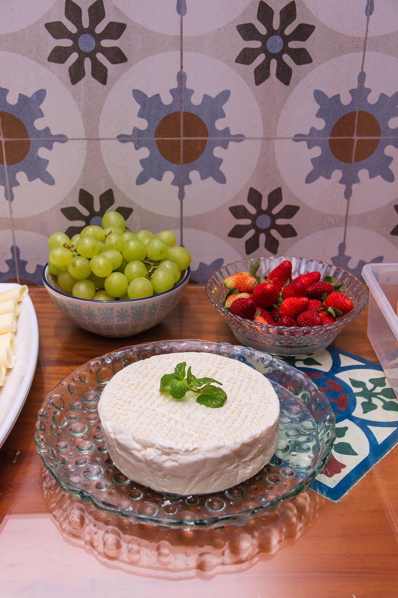 Una mesa con un bol de uvas verdes, un bol de fresas y un queso redondo sobre un plato de vidrio.