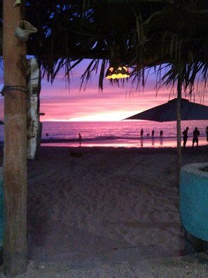 Escena de playa con atardecer, palmeras y gente caminando.
