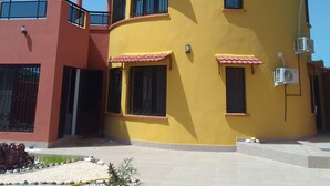 Un edificio amarillo de varios pisos con balcones, unidades de aire acondicionado y un pequeño jardín con piedras decorativas y plantas.