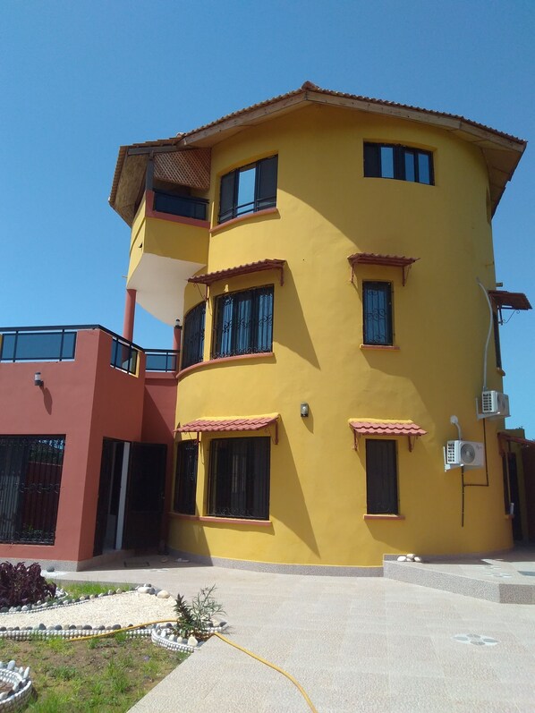 Un edificio amarillo de varios pisos con balcones, unidades de aire acondicionado y un pequeño jardín con piedras decorativas y plantas.