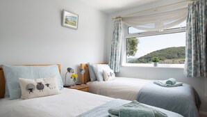 A bedroom with two beds, a nightstand with a lamp, a window with curtains, and a view of the outdoors. - Heddfan (Conwy)
