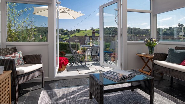 A sunlit room with a glass door leading to a patio, wicker furniture, and a view of greenery and hills. - Heddfan (Conwy)