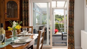 A dining area with a wooden table set for two, a cabinet with glass doors, and a view of a garden through a glass door. - Heddfan (Conwy)