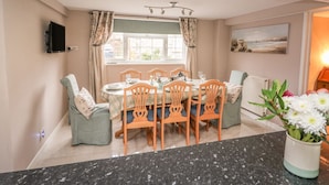 A dining area with a table set for four, surrounded by wooden chairs with blue cushions. A window with curtains is visible, and a painting hangs on the wall. - Fisher Granary (Lampeter)