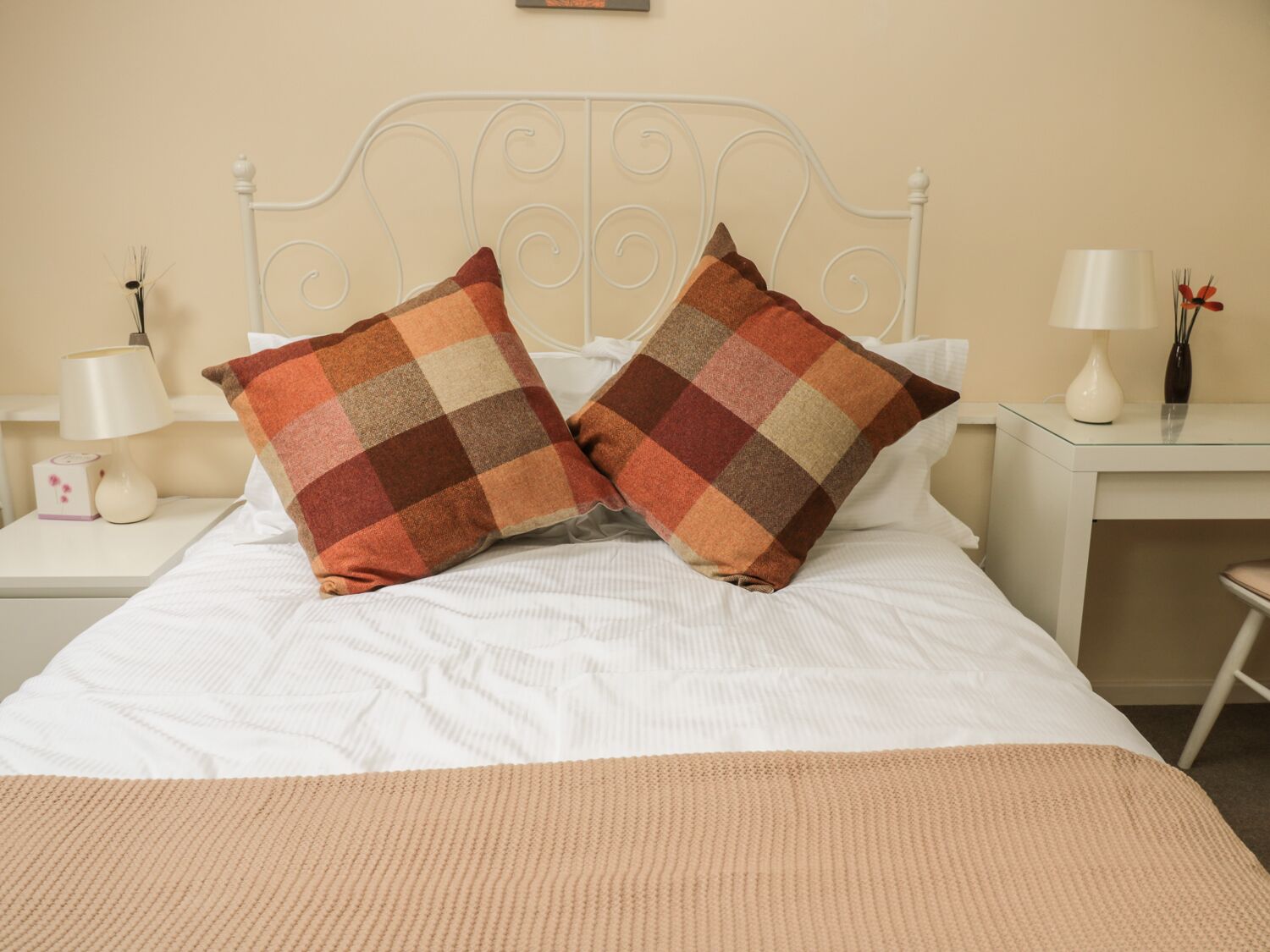 A neatly made bed with two checkered pillows, a bedside table with a lamp, and a small vase with flowers.