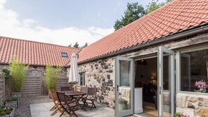 Un bâtiment en pierre avec un toit en tuiles, une terrasse avec une table et des chaises, et une porte-fenêtre coulissante donnant sur un espace intérieur.